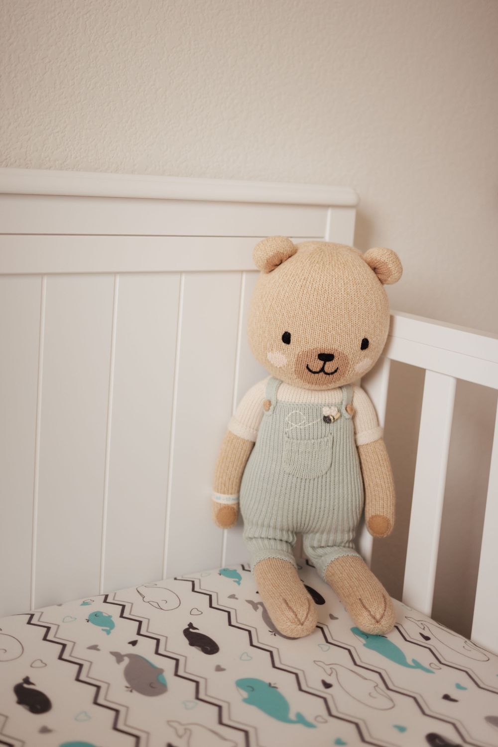 A beige stuffed teddy bear wearing light gray overalls sits propped against a white crib railing.
