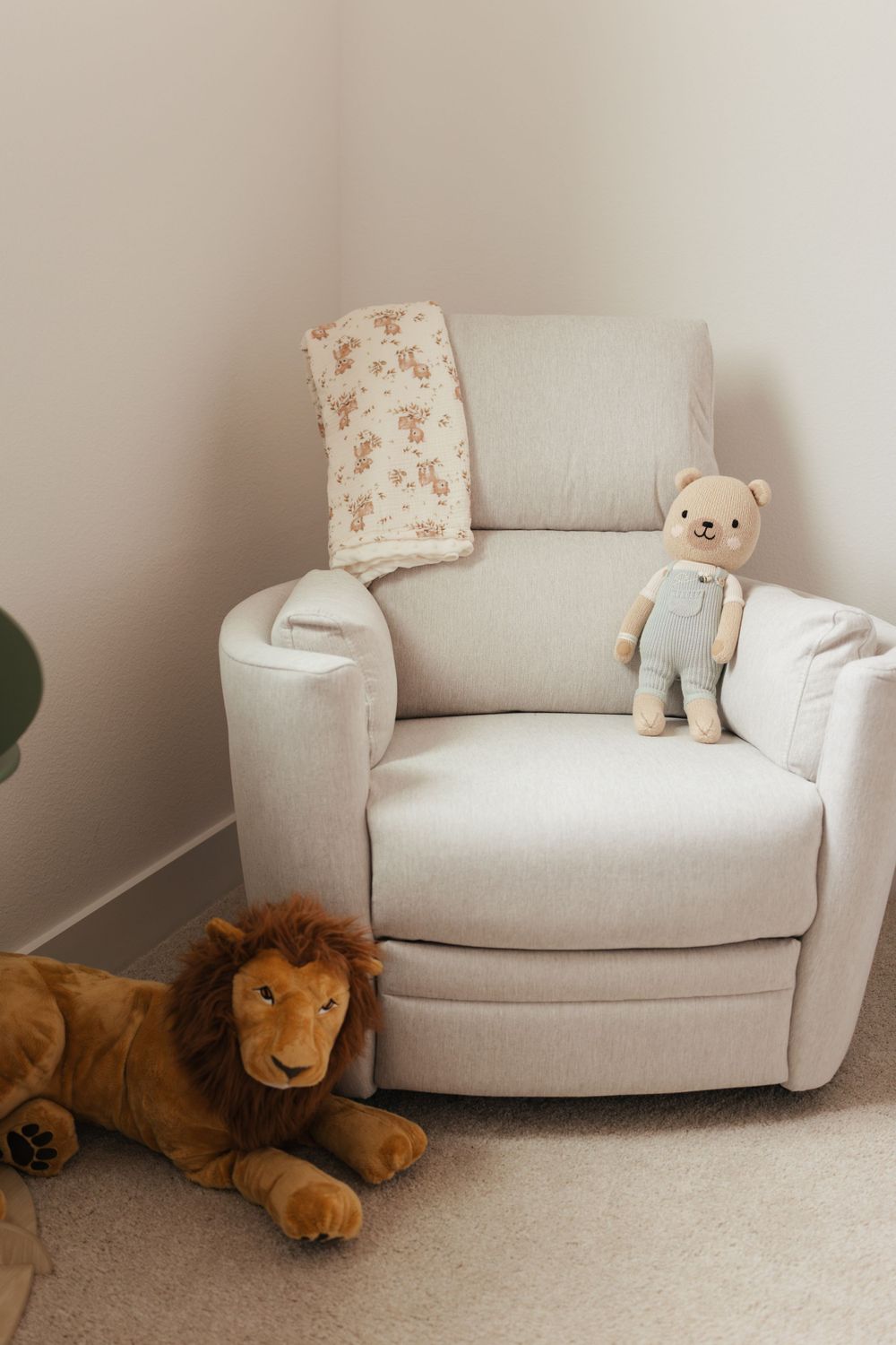 Cozy beige nursery recliner with plush lion toy on floor and teddy bear in overalls on the chair.