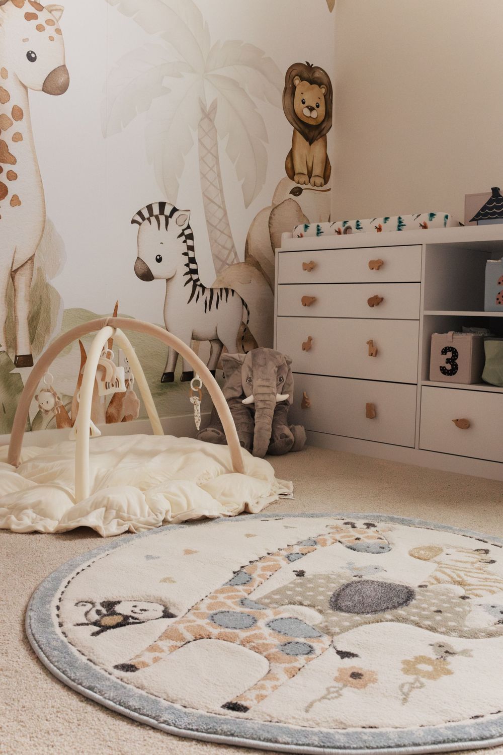Neutral safari-themed nursery with play mat, activity gym, dresser and animal wall decals in beige and gray tones.