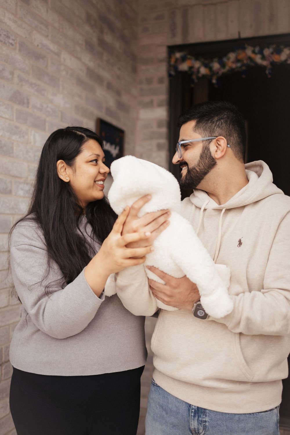 A couple holding and gazing adoringly at their swaddled baby in white blankets while standing in a cozy indoor setting.