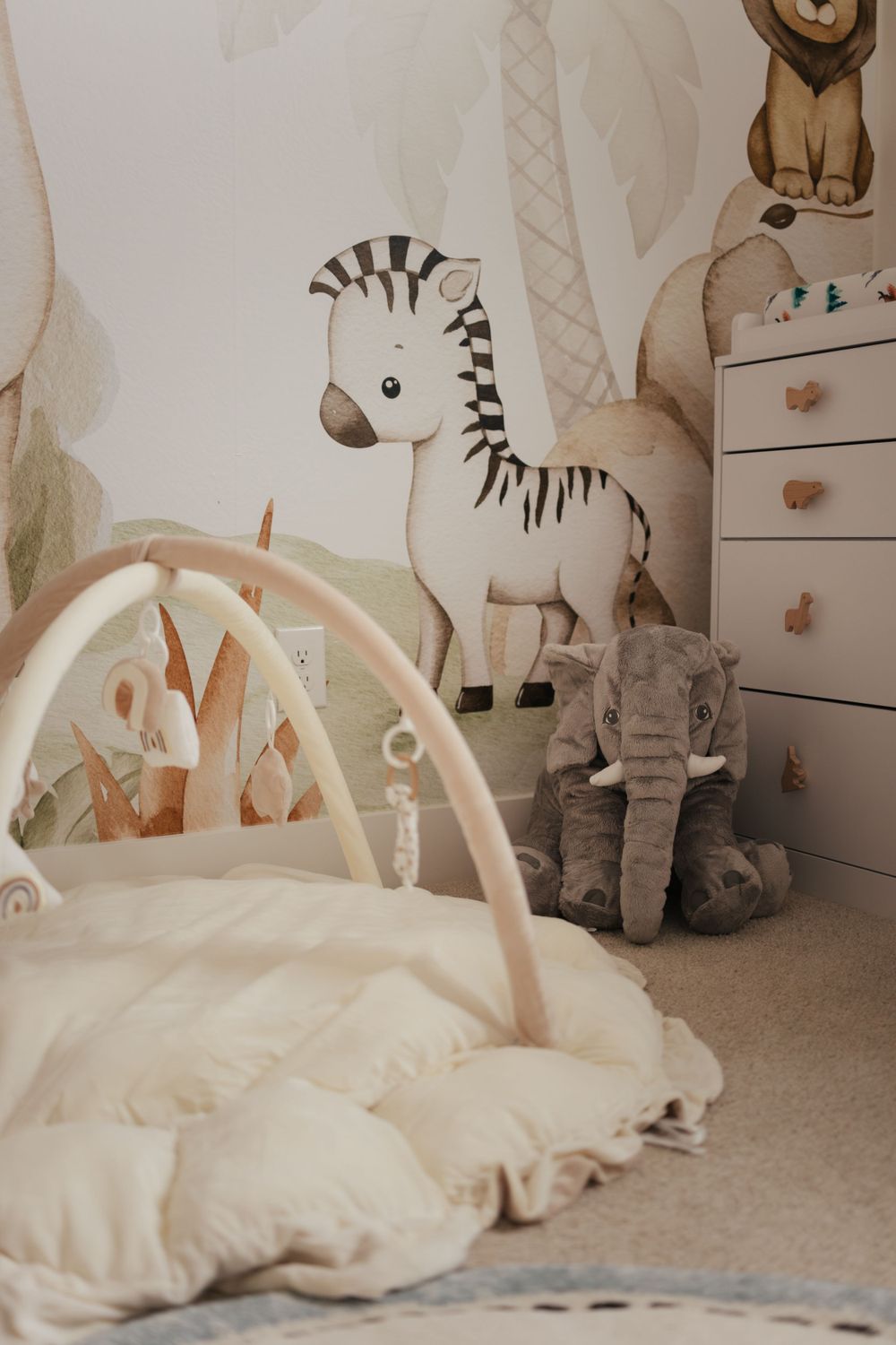 A cozy nursery corner with a plush play mat, stuffed zebra toy, elephant plush and white dresser in neutral tones.