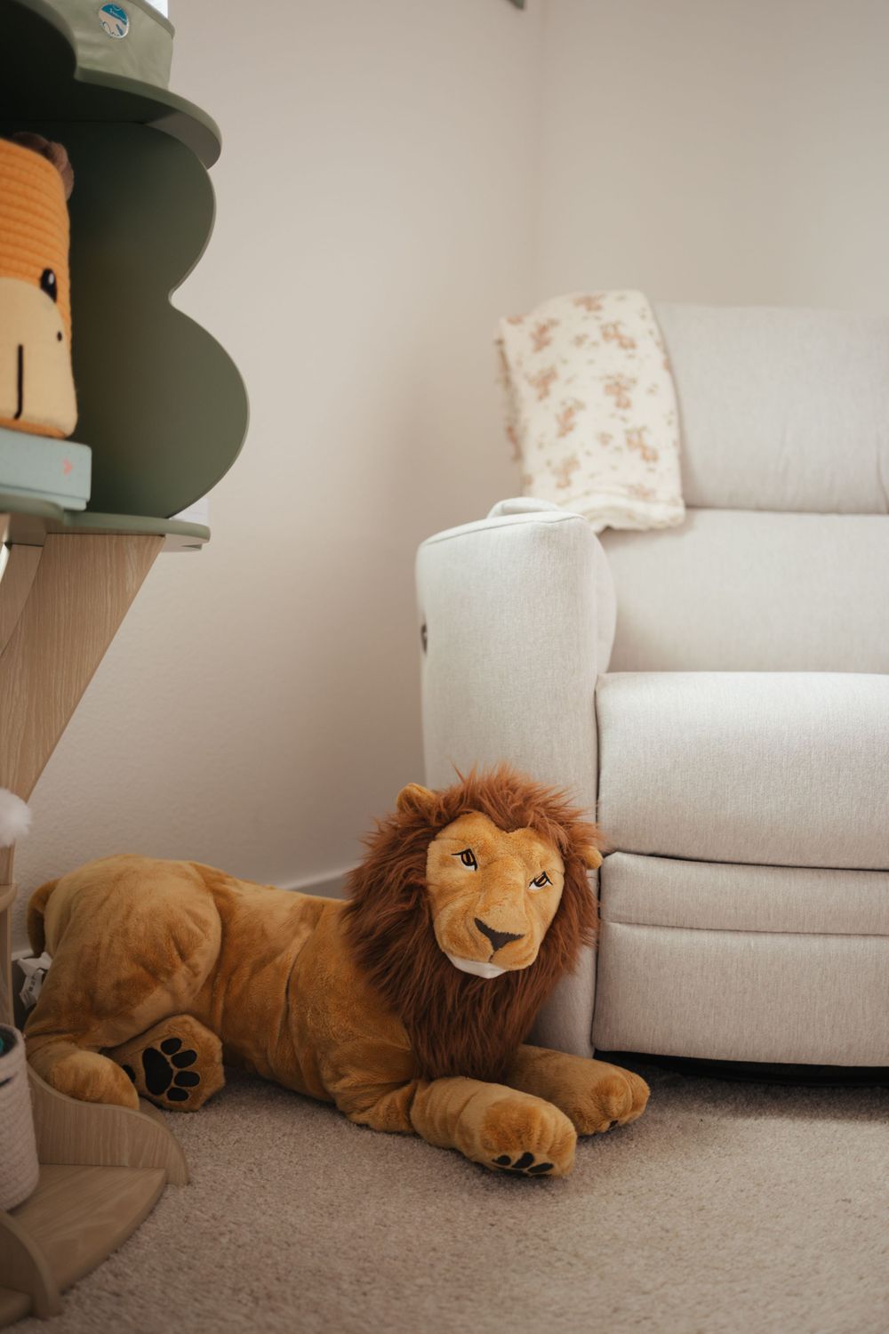 A large plush lion stuffed animal rests beside a light gray couch in a nursery with floating shelves.
