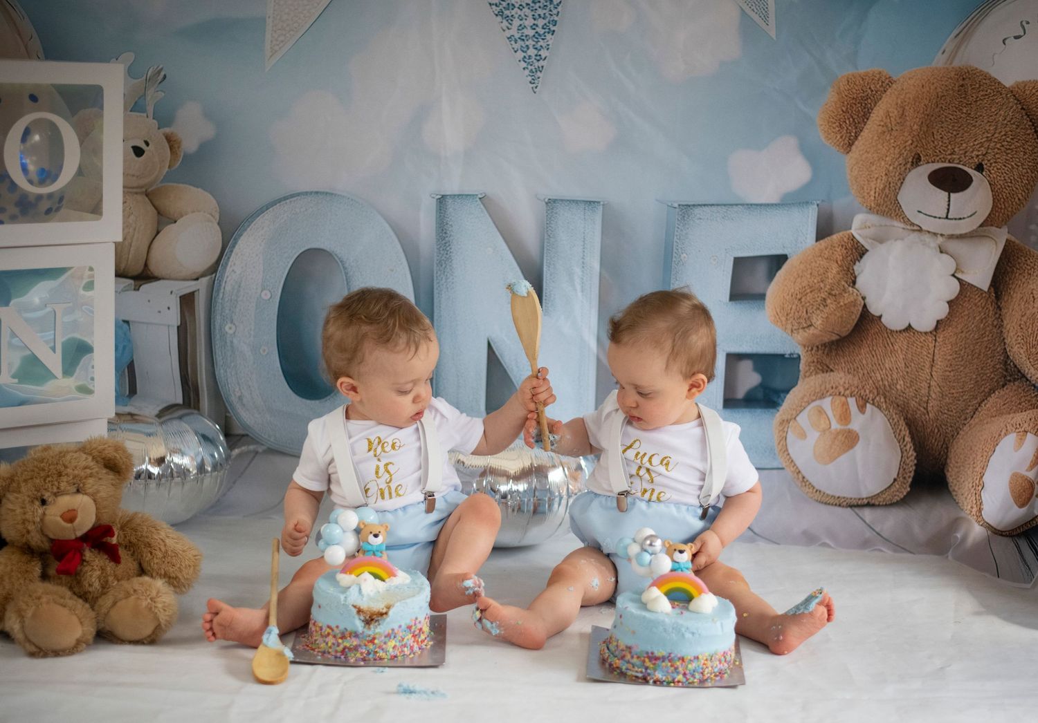 Two babies in matching blue outfits sit on the floor with stuffed bears and ONE letter decor for their first birthday celebration.