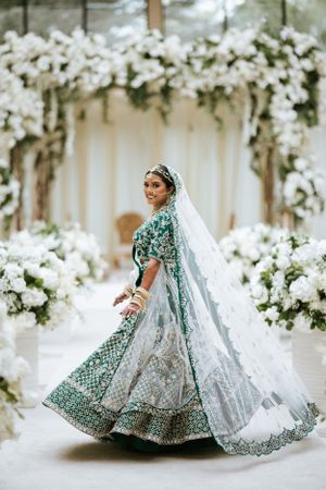 Elegant South Asian bride in green lehenga walks the aisle at Lucien’s Manor, surrounded by lush white floral wedding decor.