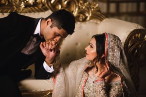 Romantic South Asian wedding moment at The Marigold in Somerset, NJ as the groom kisses the bride’s hand on a regal gold sofa.