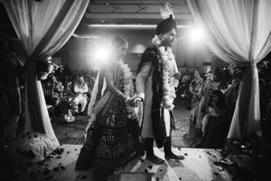 Black and white photo of a South Asian wedding ceremony with the couple taking pheras under a floral mandap surrounded by family and friends