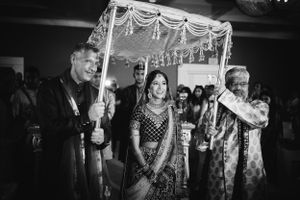 Black and white photo of a South Asian bride entering under a floral canopy, surrounded by family during her wedding in New Jersey.