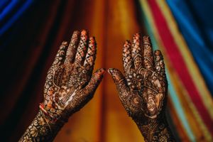 Close-up of bridal mehndi designs on both hands, featuring intricate patterns against a vibrant, colorful backdrop at a South Asian wedding.