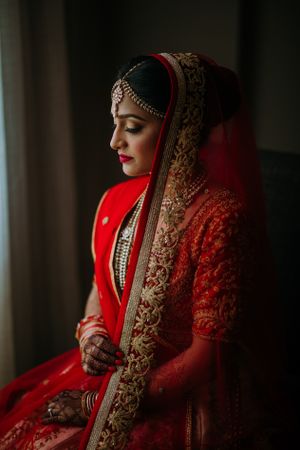 South Asian bride in red lehenga sits by a window at Falls Church Marriott Fairview Par, captured in soft, natural light before the ceremony