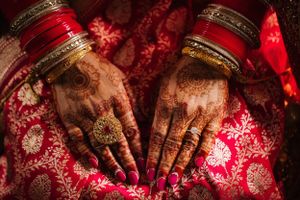 Close-up of South Asian bride’s hands with intricate mehndi, red bangles, and gold jewelry on embroidered bridal lehenga.