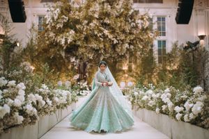 South Asian bride in pastel lehenga walking down a floral aisle at Casa Cipriani New York, surrounded by elegant white and gold décor.