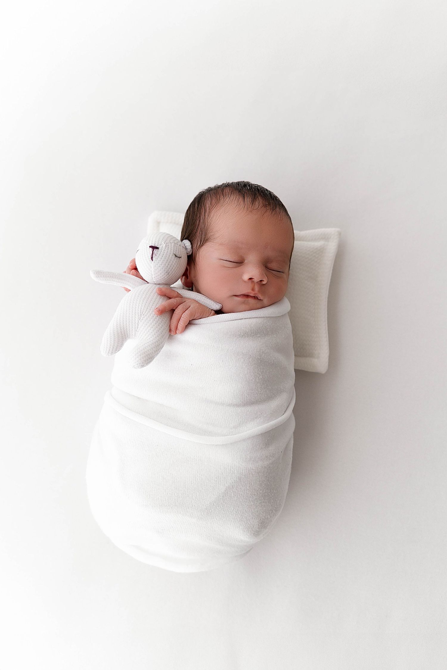 Peaceful sleeping newborn wrapped in white blanket with small plush toy.