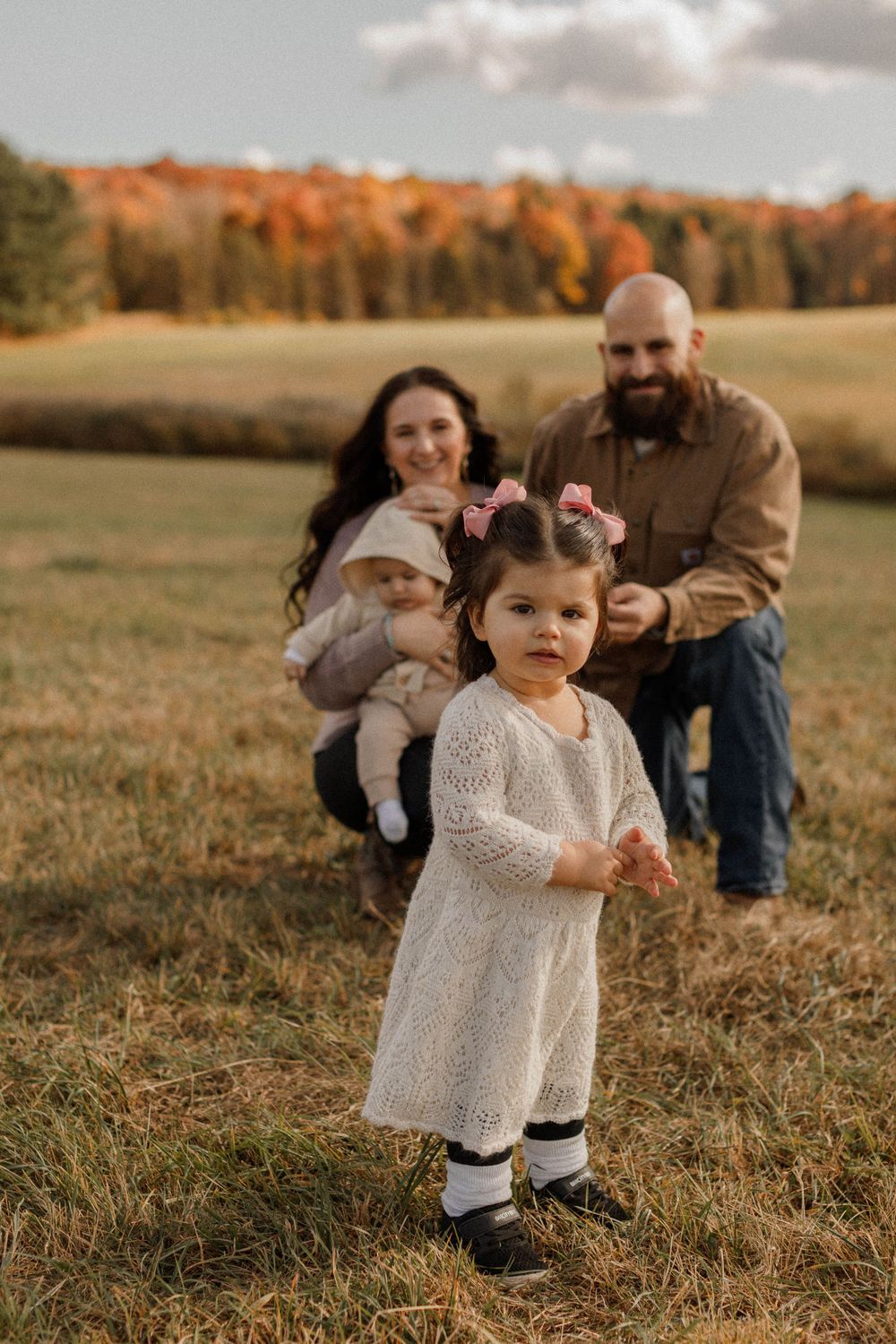 Family portrait in autumn field with small child in white dress in foreground.