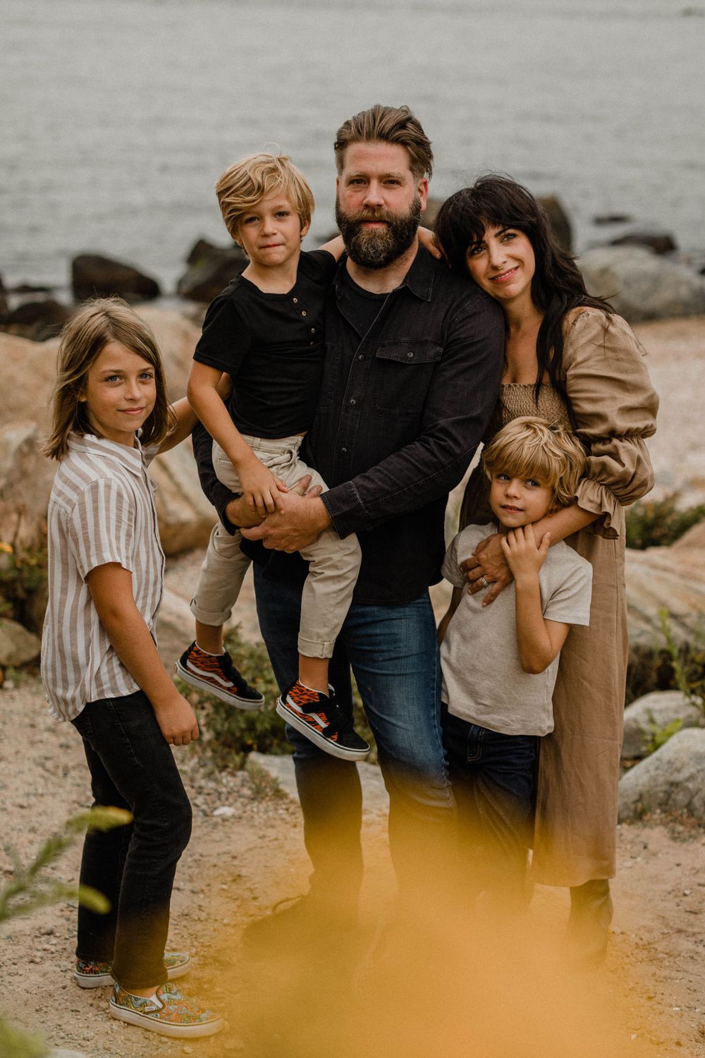A family of five poses together on rocky coastal shore at sunset.