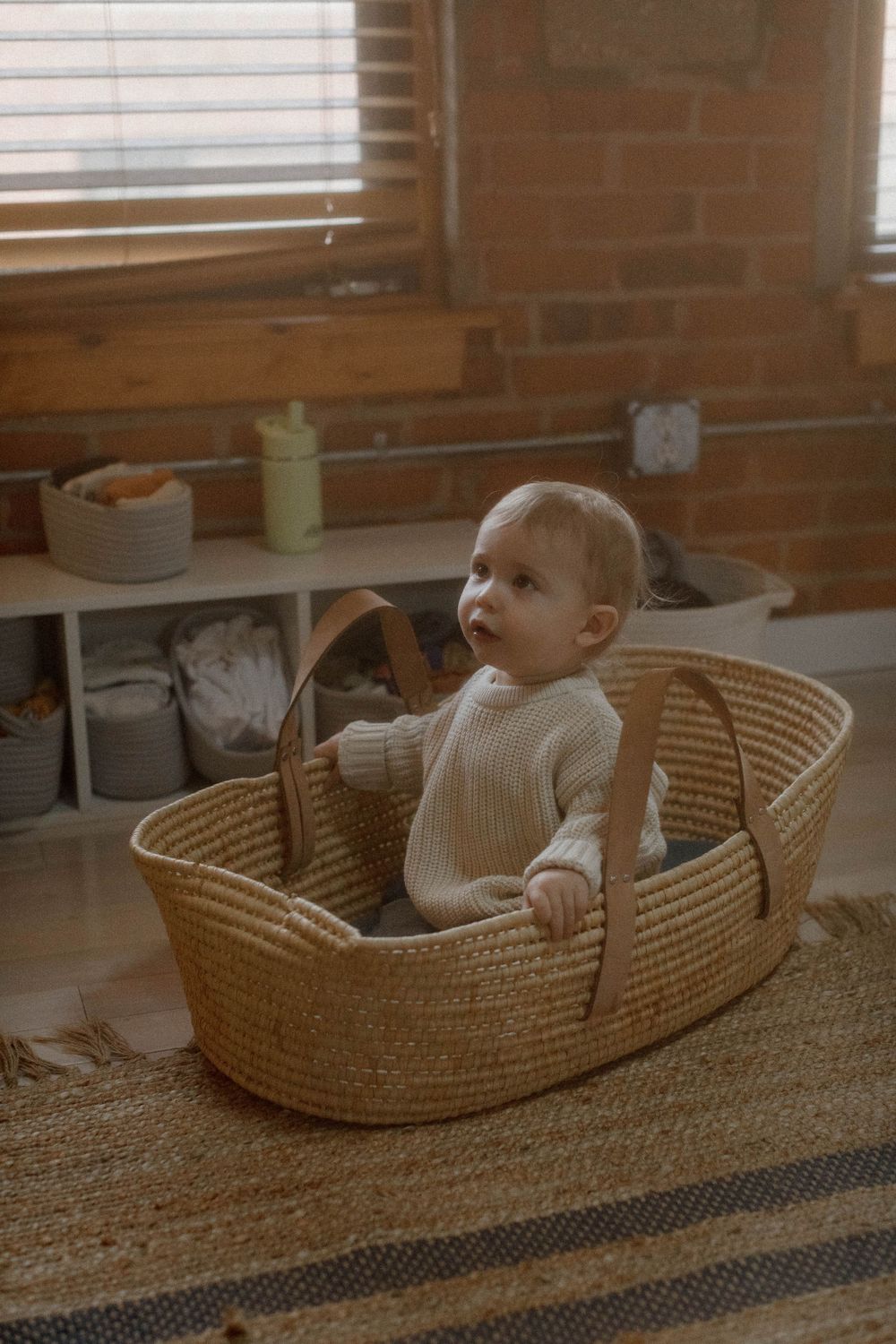 Baby in white sweater sits inside woven basket on textured rug near brick wall.