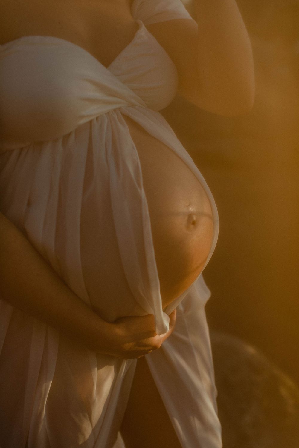 Artistic close-up of pregnant belly draped in white fabric with golden light.