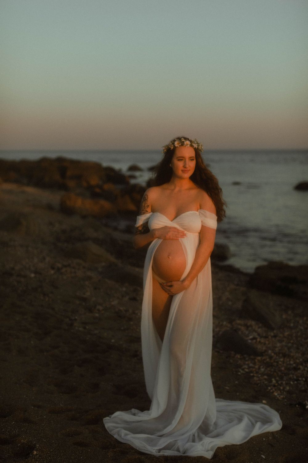 Expectant mother in flowing white dress poses at sunset on rocky coastline.