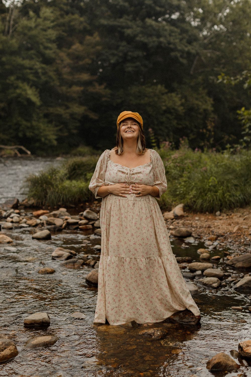A woman in a floral dress stands in a shallow rocky stream wearing an orange cap.