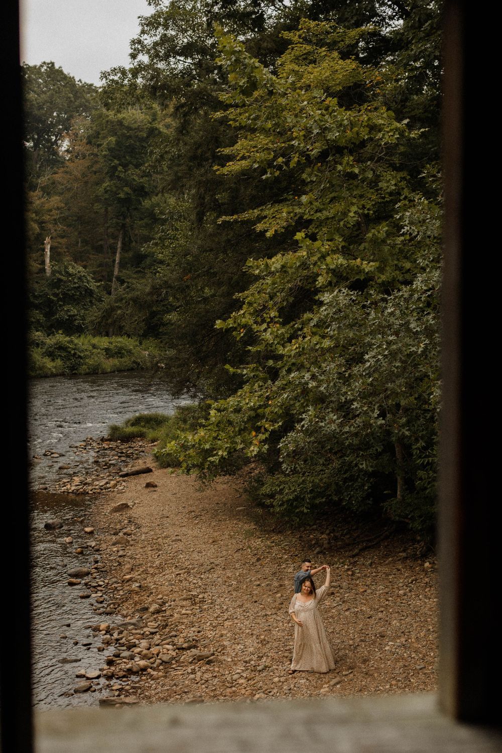 A person in a flowing dress poses by a tree-lined riverbank.