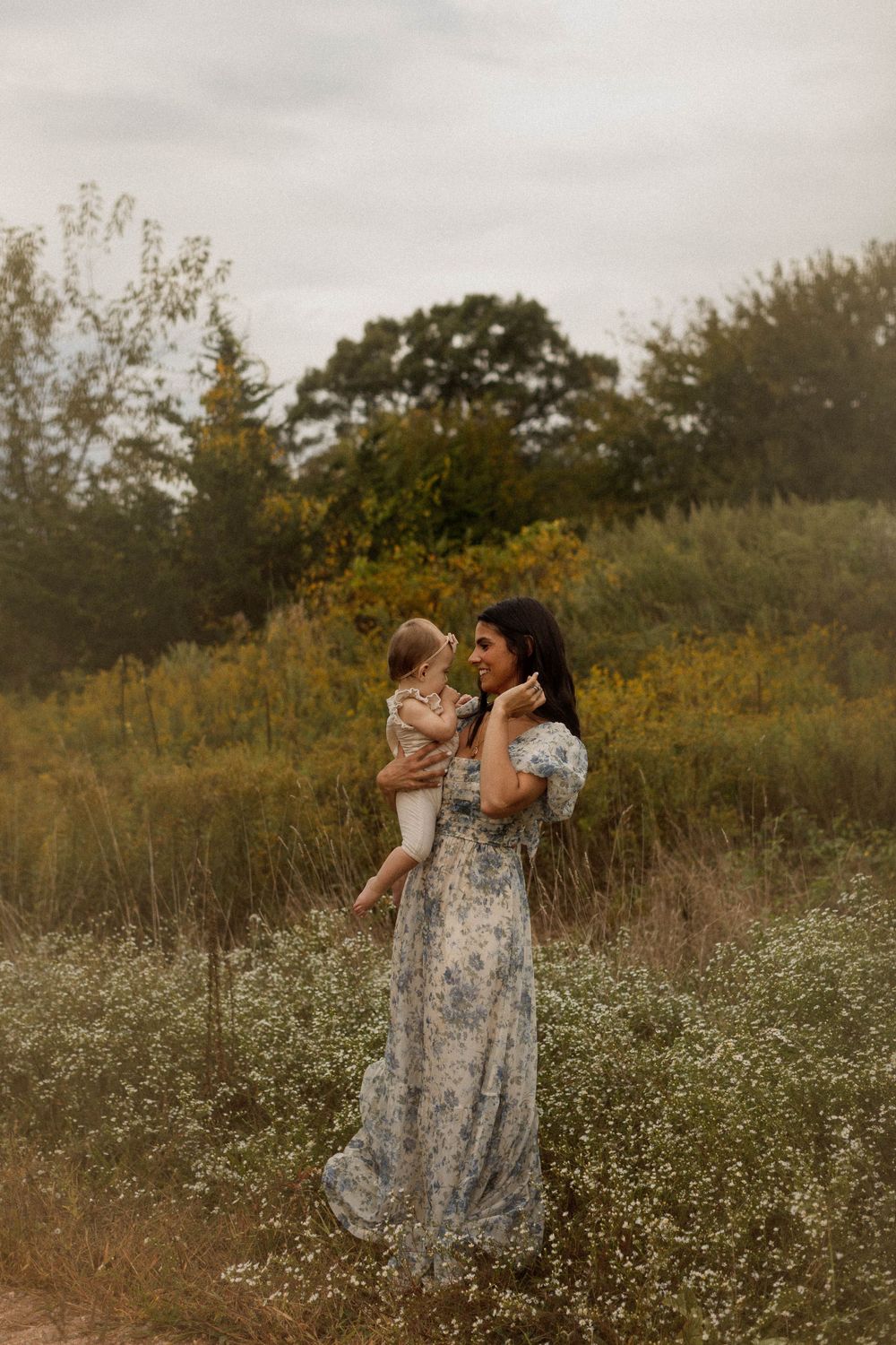 A woman in a floral dress holds a small child in a field of wildflowers at dusk.
