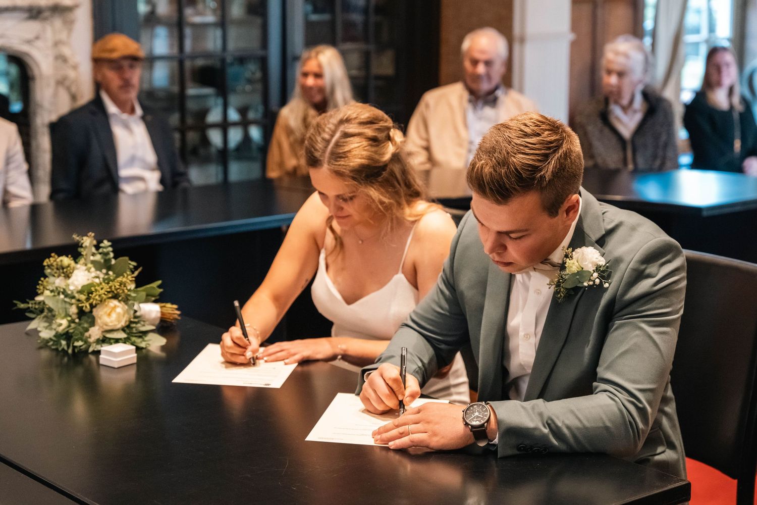 A couple signing marriage documents at a dark table with floral decorations.