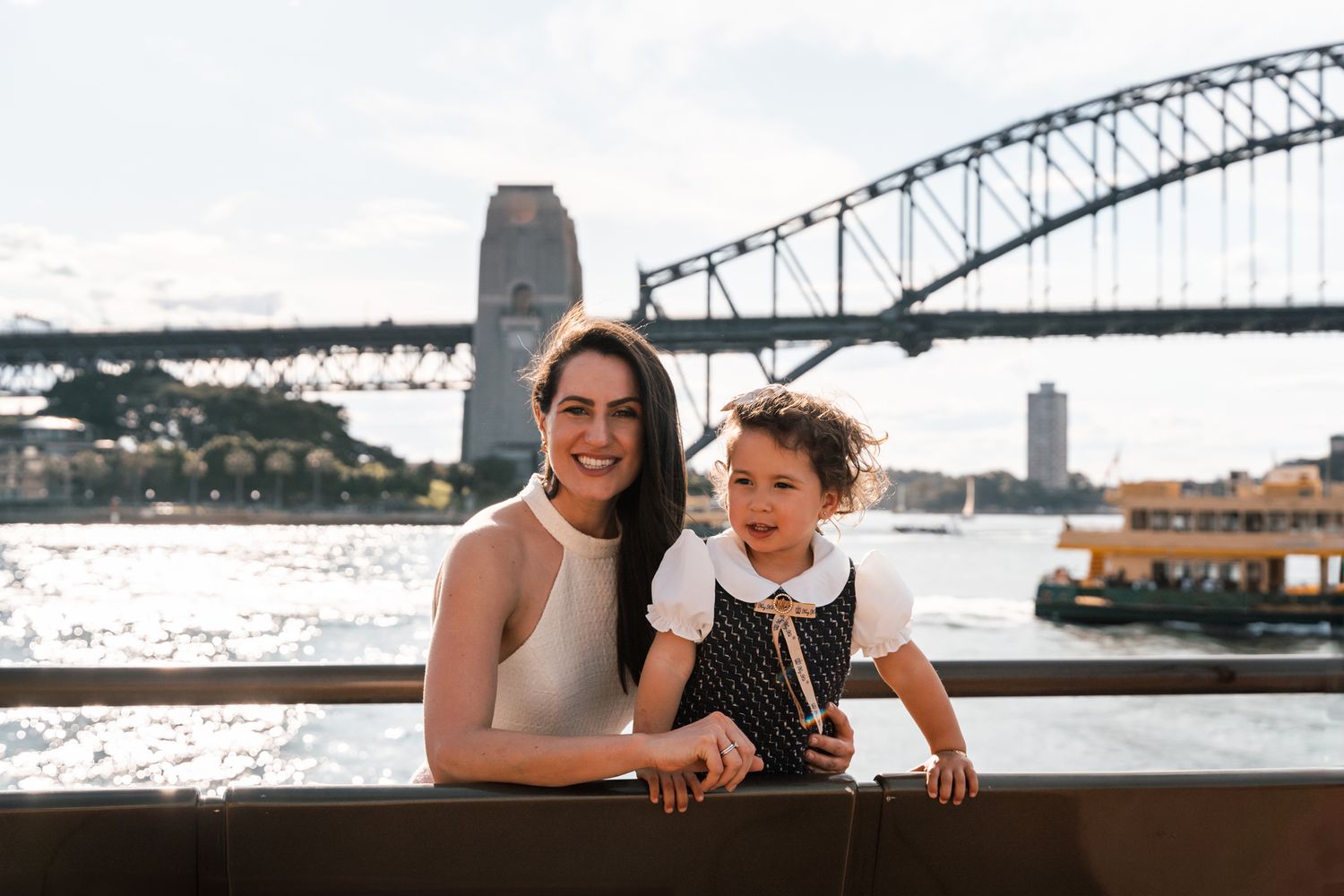 A mom and daughter share a joyful smile during a Sydney family photoshoot, with the Harbour Bridge and Sydney Ferry