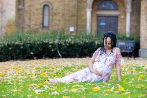 A pregnant woman in a floral dress relaxes on autumn grass with fallen yellow leaves near a stone building.