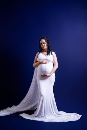 Maternity photo in white flowing gown against deep blue studio background.