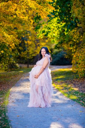 A woman in a flowing pink gown poses on a path surrounded by autumn foliage.