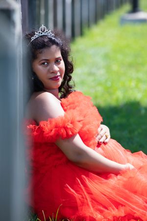 Maternity portrait with woman in red tulle dress and tiara sitting outdoors.