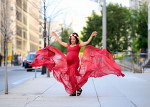 Woman in flowing red dress twirls on city sidewalk during maternity photoshoot.