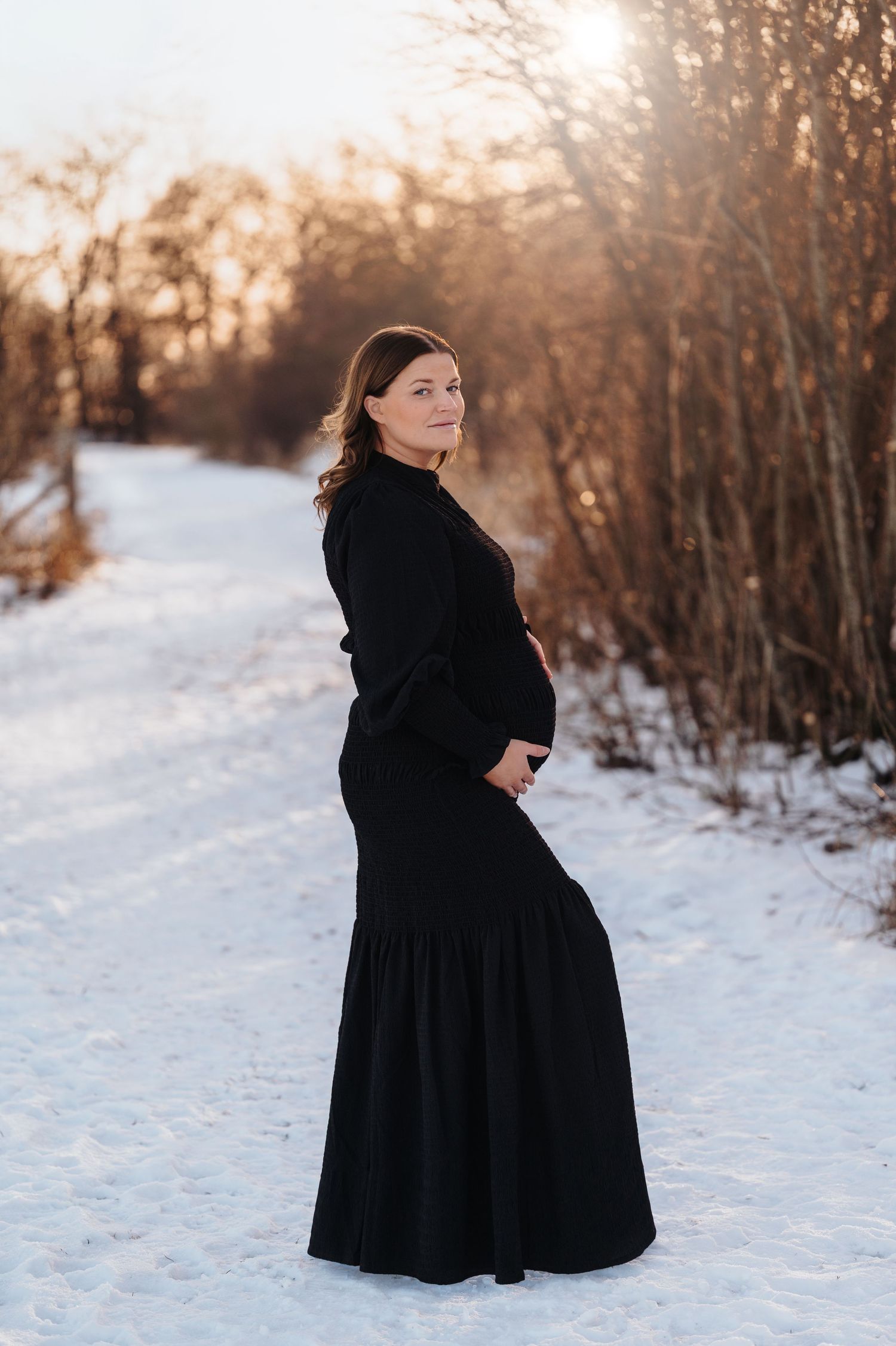 Elegant maternity portrait featuring a long black dress in a snow-covered winter setting.