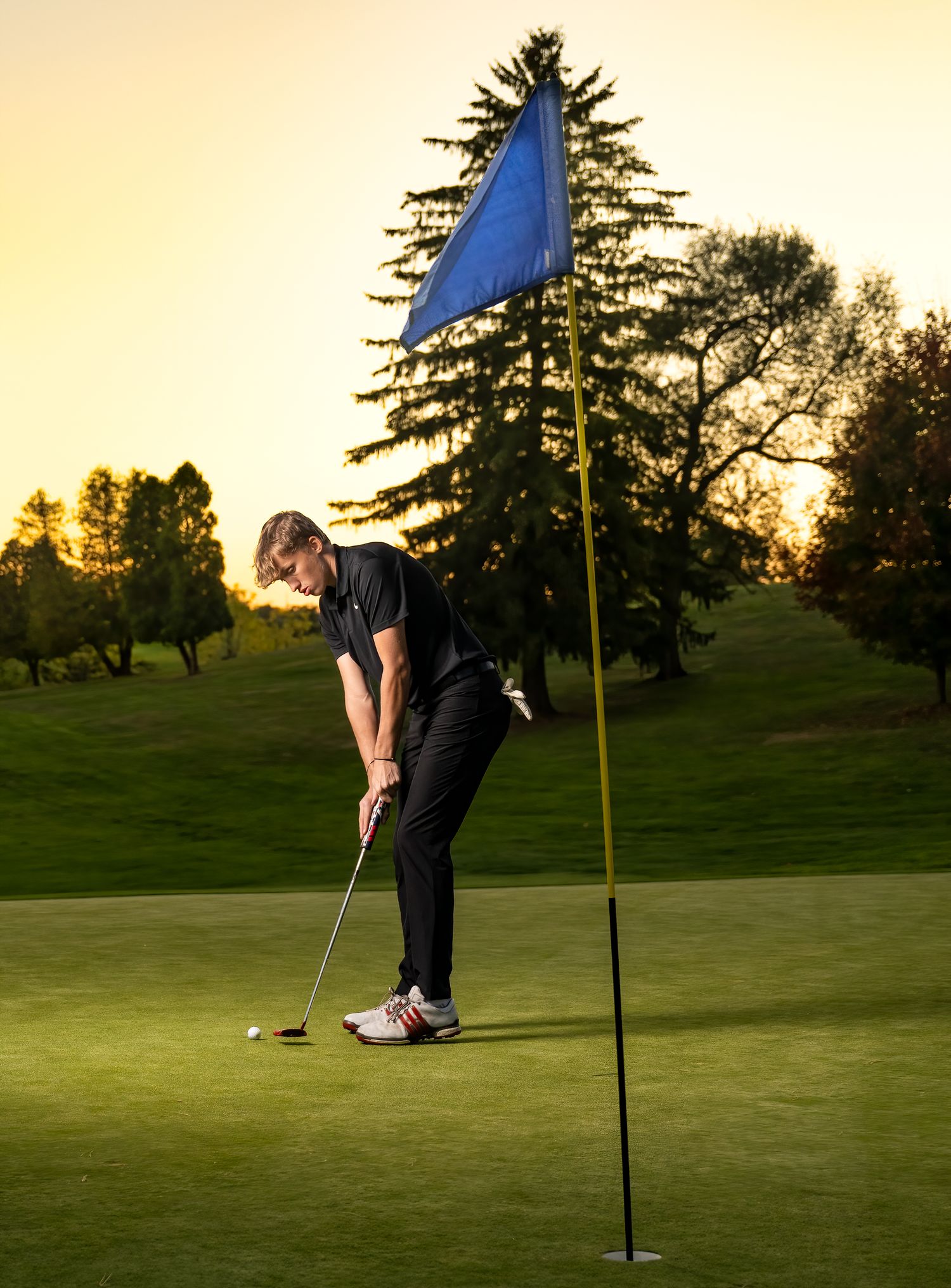 Golfer lines up putt on green during sunset with blue flag in background.