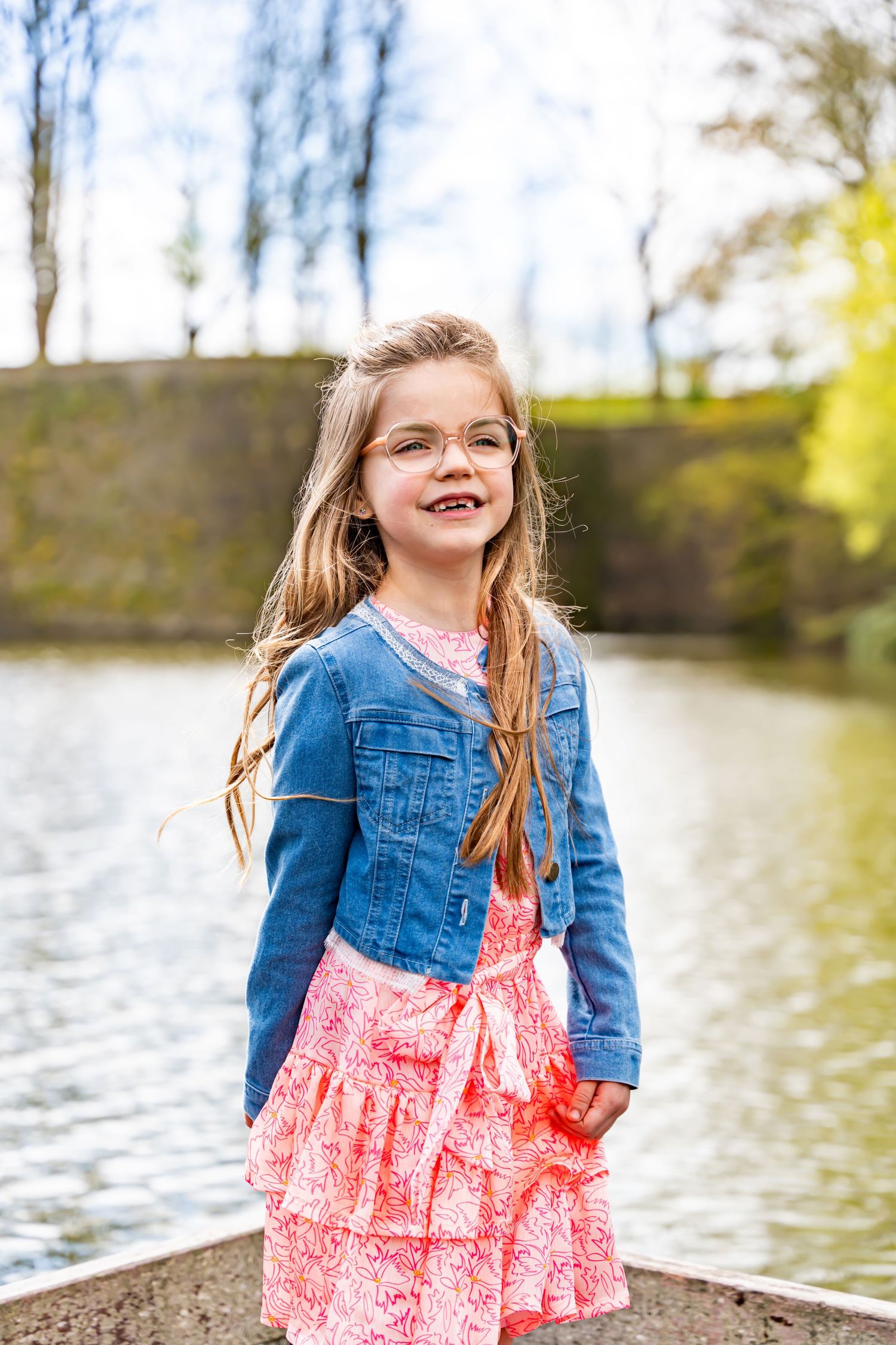 A young child wearing a denim jacket and pink floral dress poses by a lake on a sunny spring day.