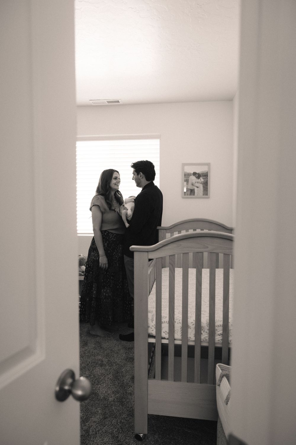Black and white photo of new parents sharing a tender moment together in a nursery by a crib.