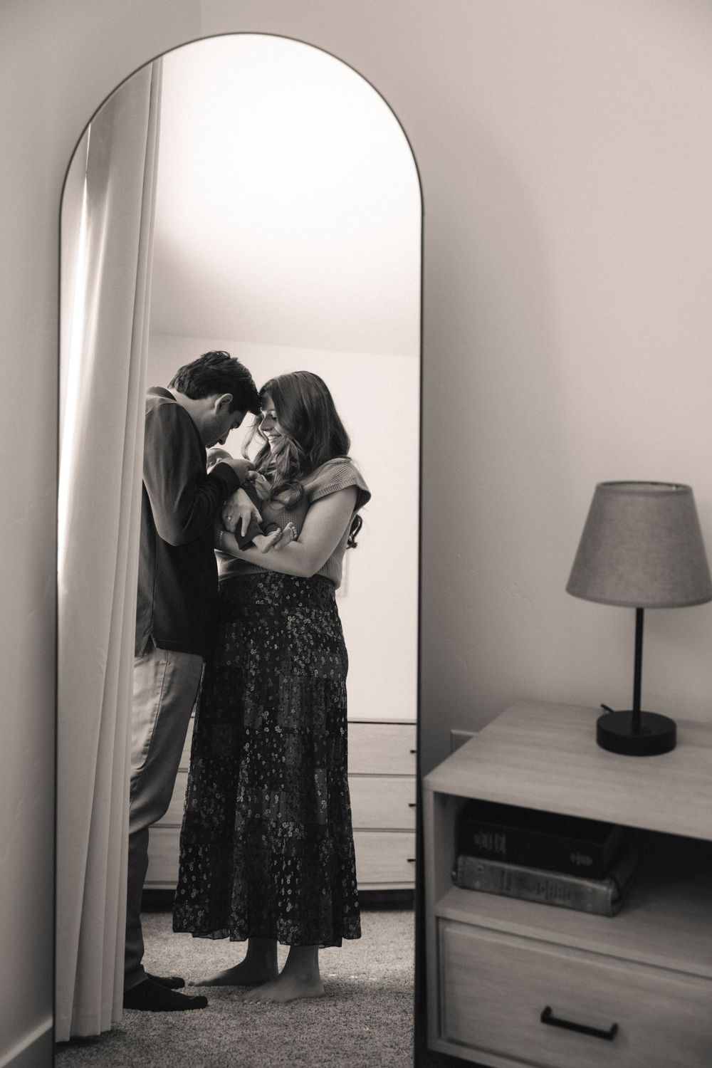 Black and white reflection of new parents kissing their newborn baby captured in a tall arched mirror.