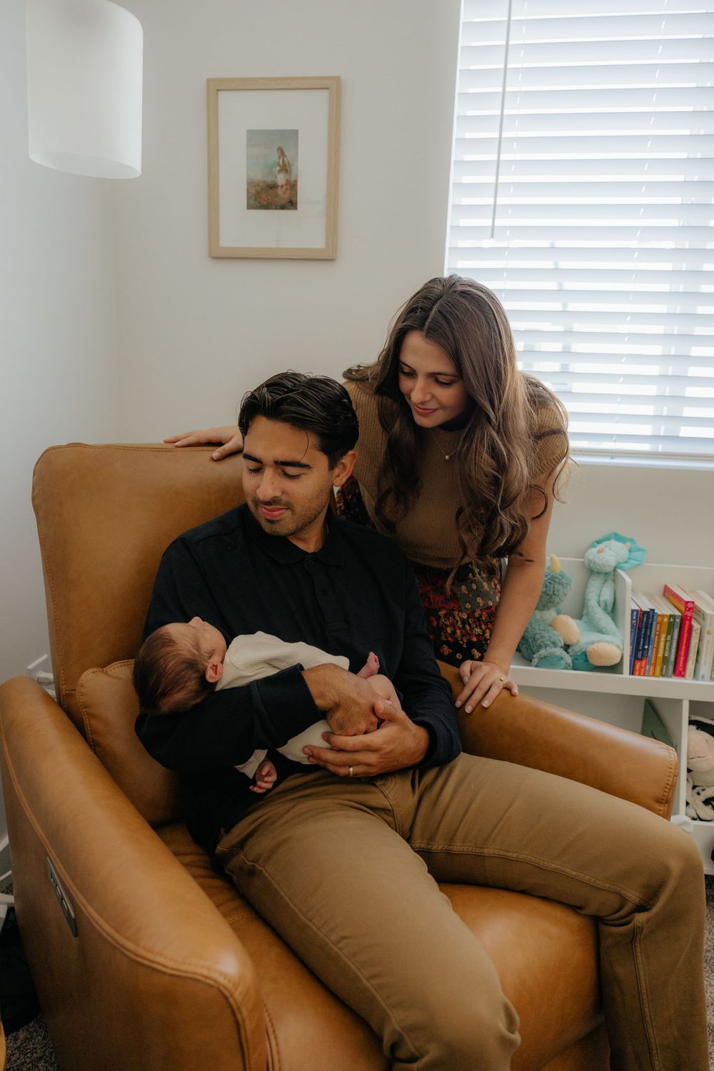 Father sitting in leather chair holding newborn while mother stands behind in a bright nursery room.