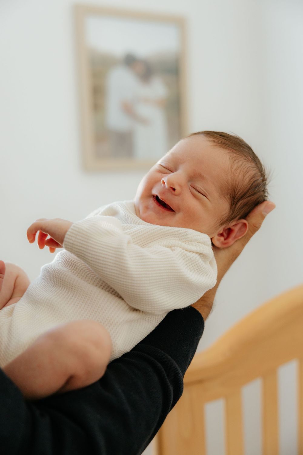 Peaceful newborn baby smiling while being gently held in a cozy white onesie near a wooden crib.