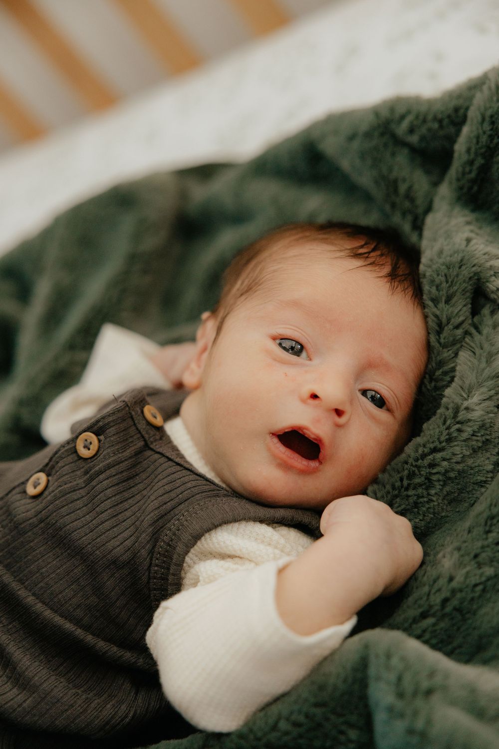 Adorable newborn baby in dark green romper lying on fluffy green blanket with eyes open and mouth agape.