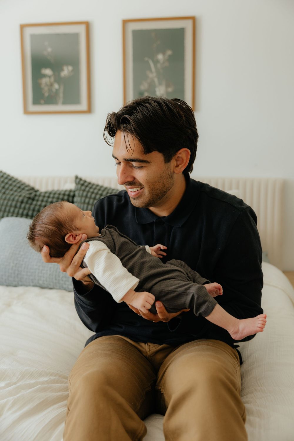 Smiling father in black shirt cradling his newborn baby while sitting on a white bed at home.