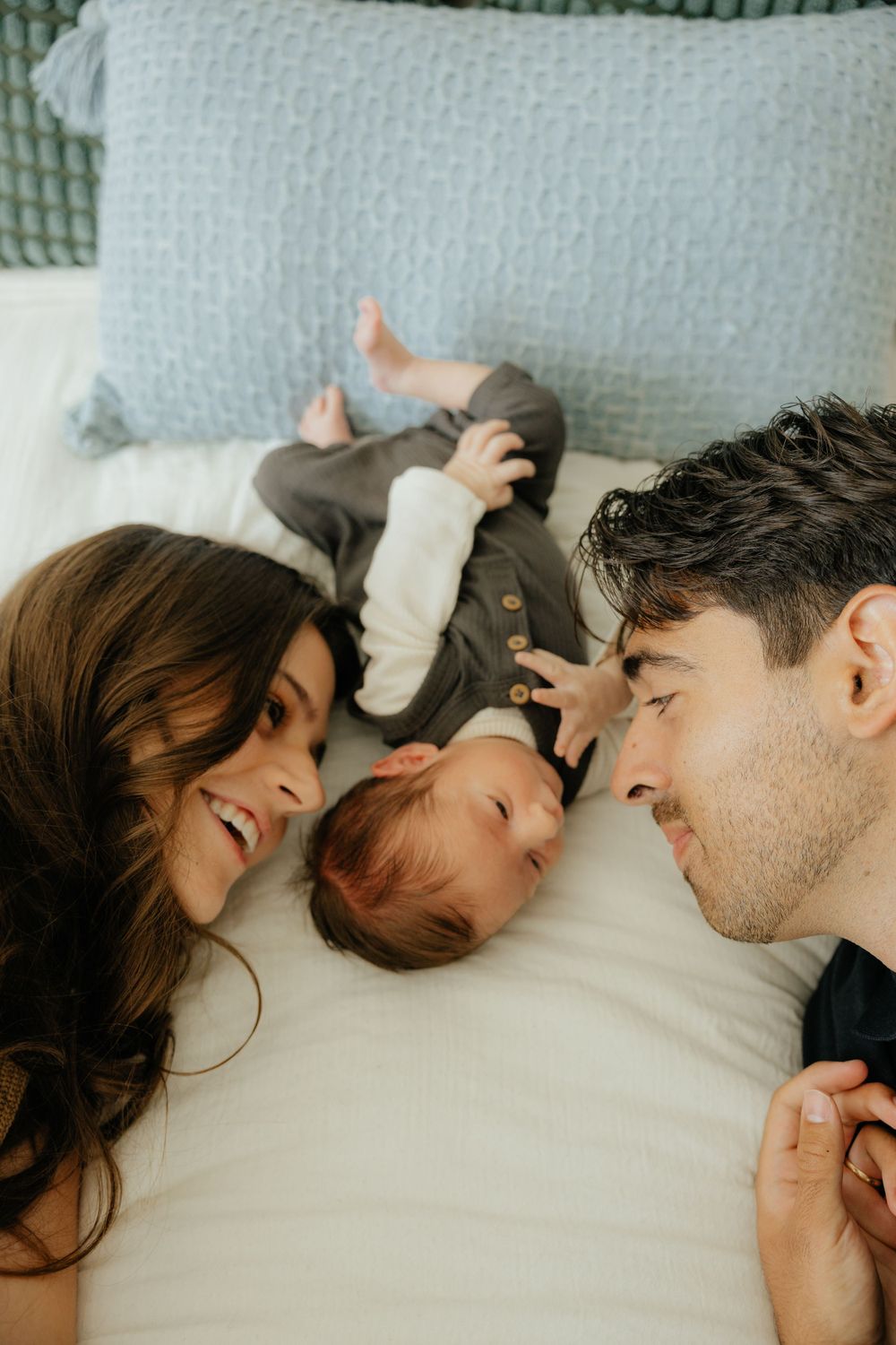 Happy new parents lying on white bed smiling at their newborn baby between them during home session.