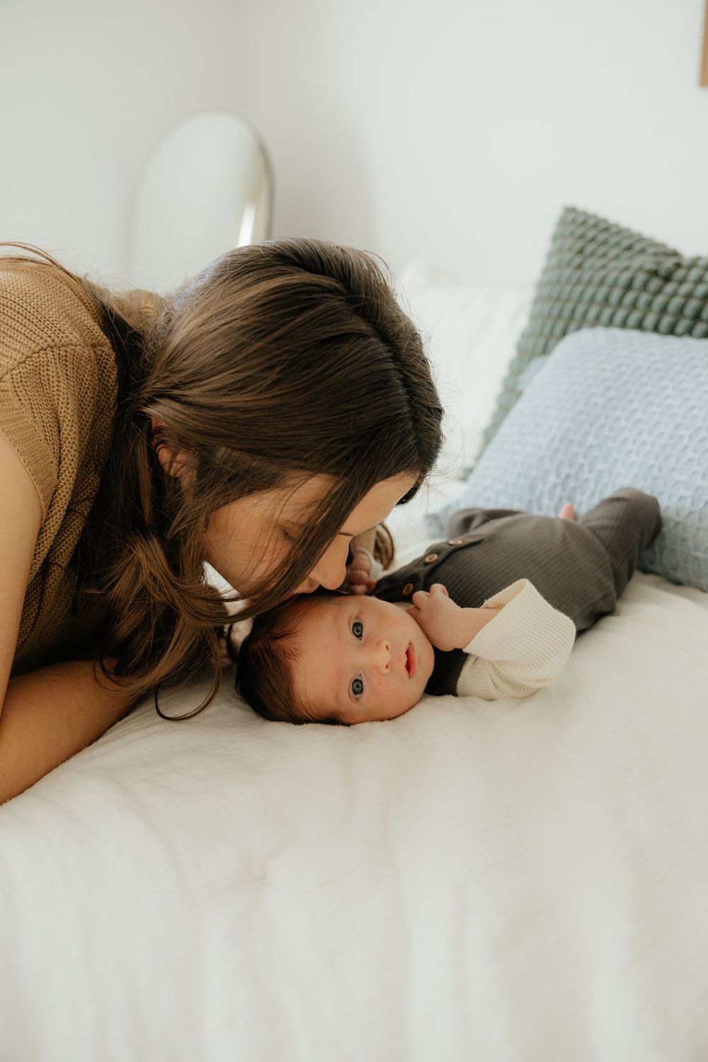 Loving mother leaning down to kiss her wide-eyed newborn baby lying on a white bed at home.