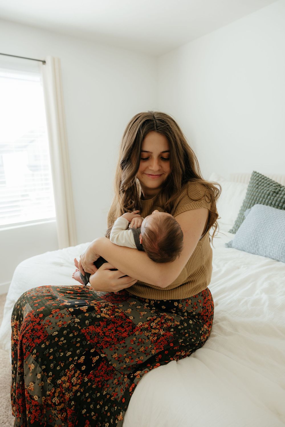 New mother in floral skirt sitting on white bed cradling and gazing at her sleeping newborn baby.