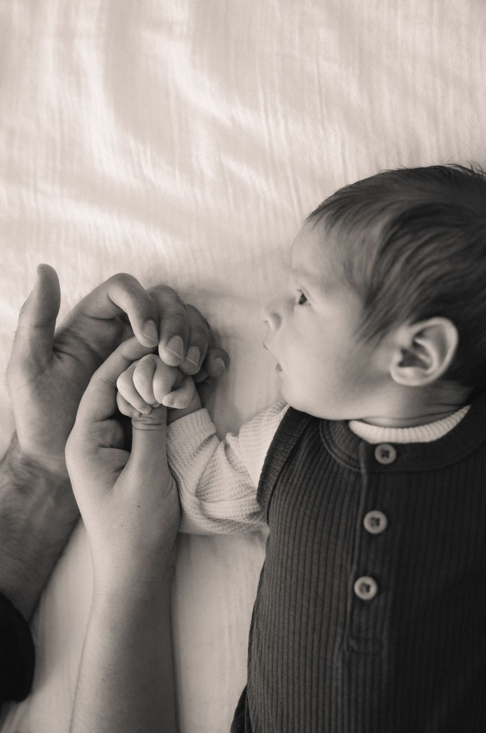 Black and white photo of newborn baby gripping parents fingers while lying on a white bed.