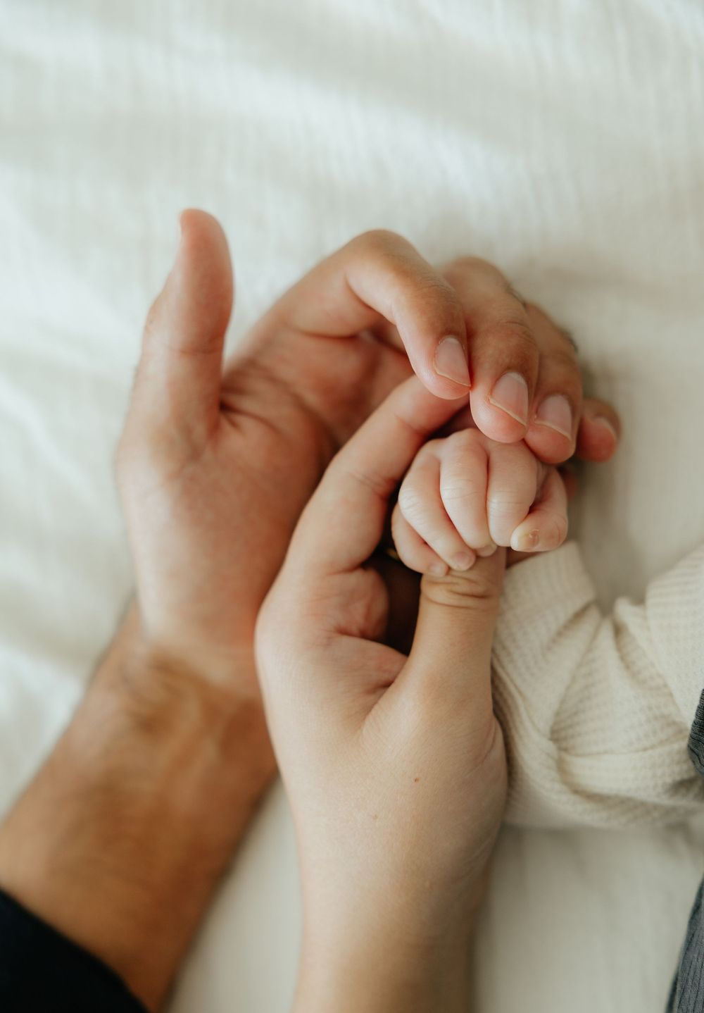 Close-up of parents gently holding their newborn baby's tiny hand on a soft white blanket.
