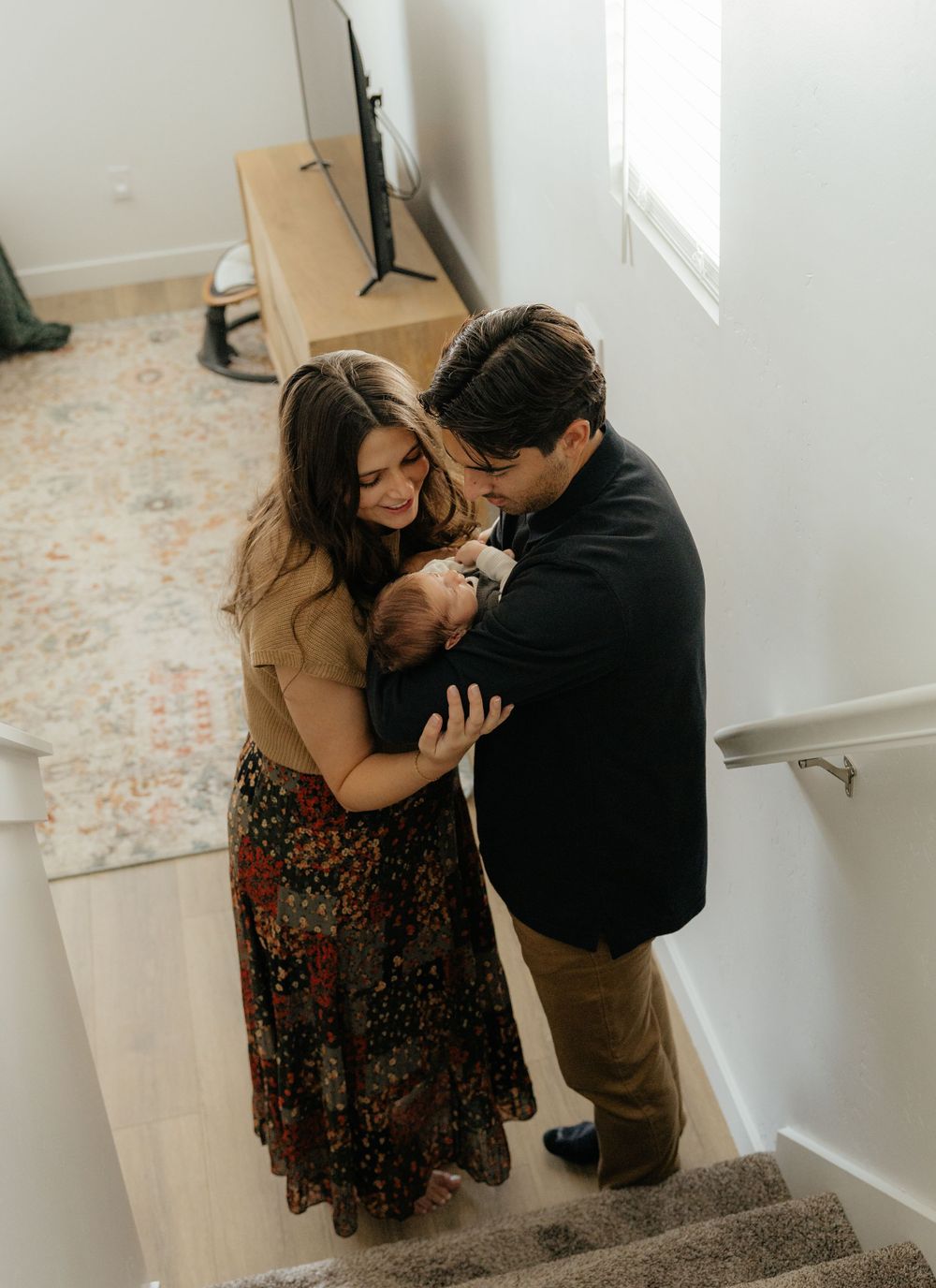 New parents stand on staircase looking down lovingly at their newborn baby in a bright modern home.