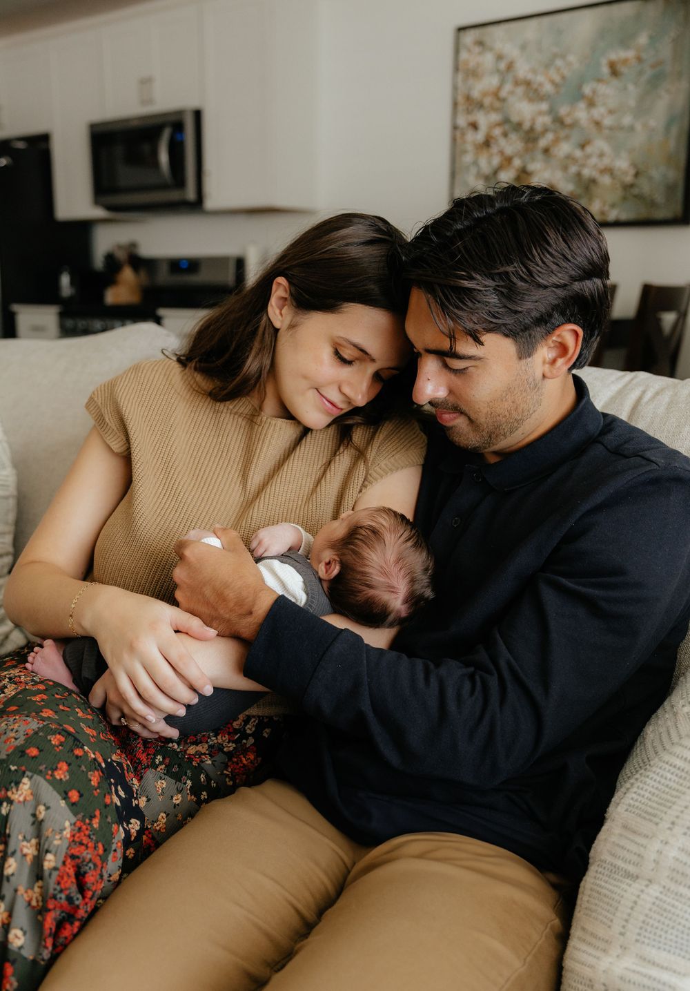 A couple sits closely on a sofa in their home, lovingly cradling their newborn baby together.
