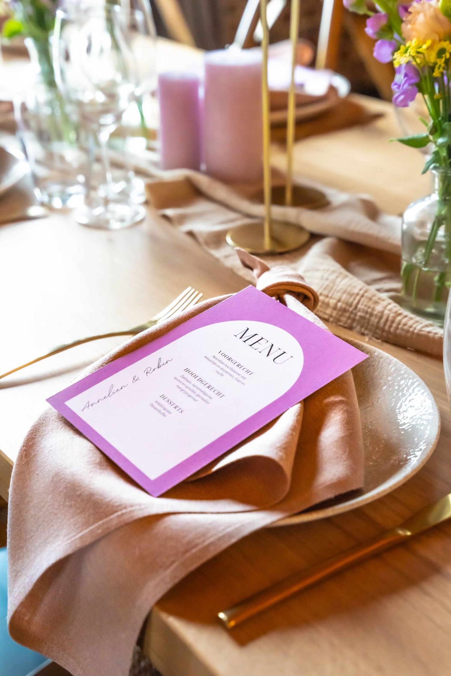 Elegant place setting with purple menu card, pink candles, and gold cutlery on beige tablecloth.