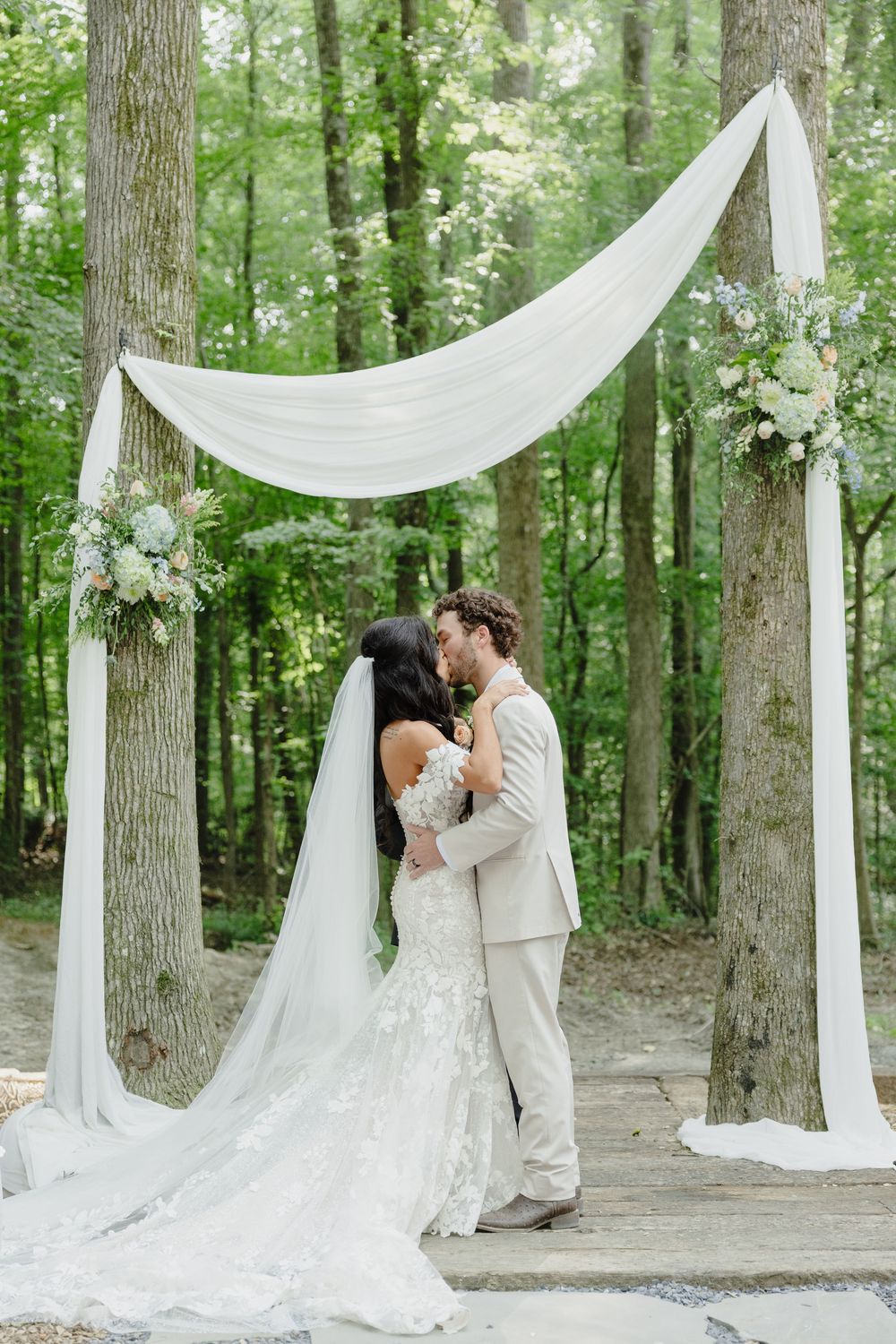 Romantic outdoor wedding ceremony in a wooded setting with flowing white drapery and floral arrangements decorating tree trunks.