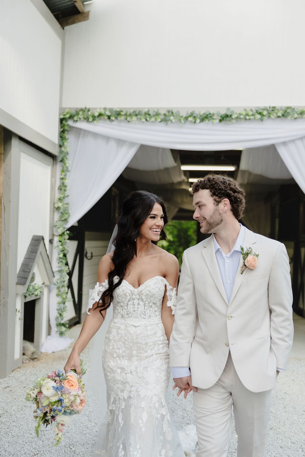 A couple in wedding attire smiles at each other while walking outdoors with floral decor and white draping in background.
